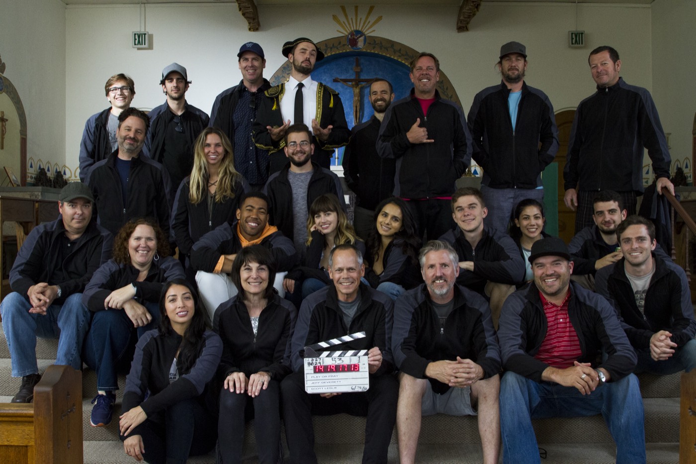 Jeff Deverett, holding the slate, front and centre with about two dozen members of the cast and crew of God Incorporated, seated on the steps of a church set